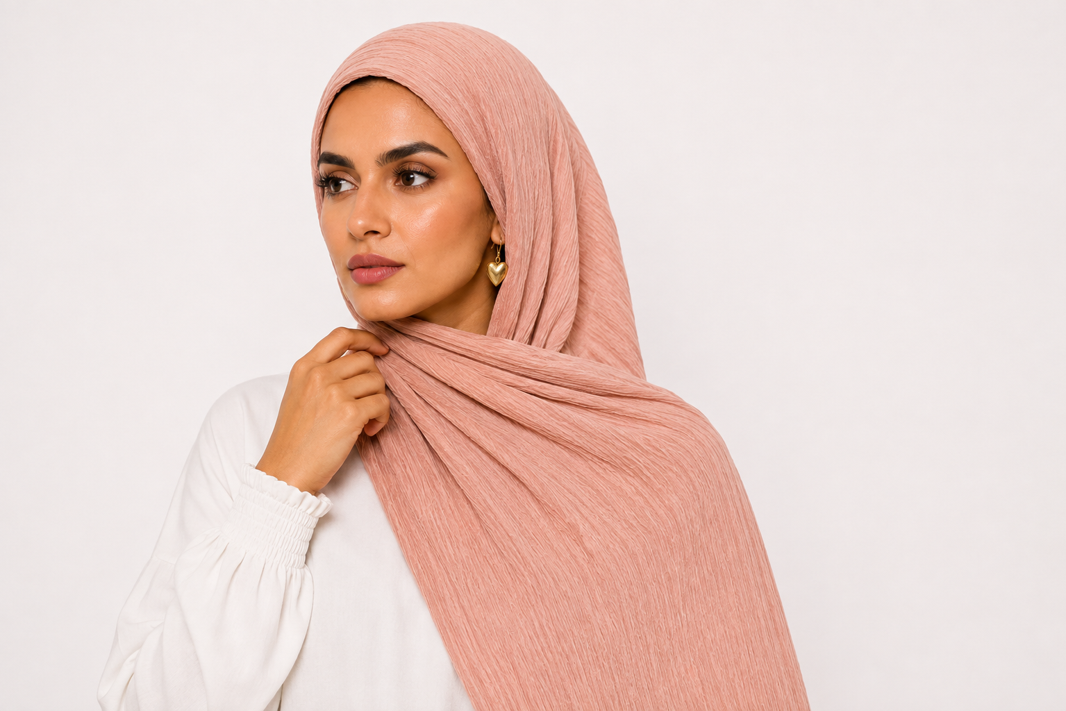 A woman wearing a dusty pink ribbed hijab and a white long-sleeve blouse, posing against a solid off-white background.