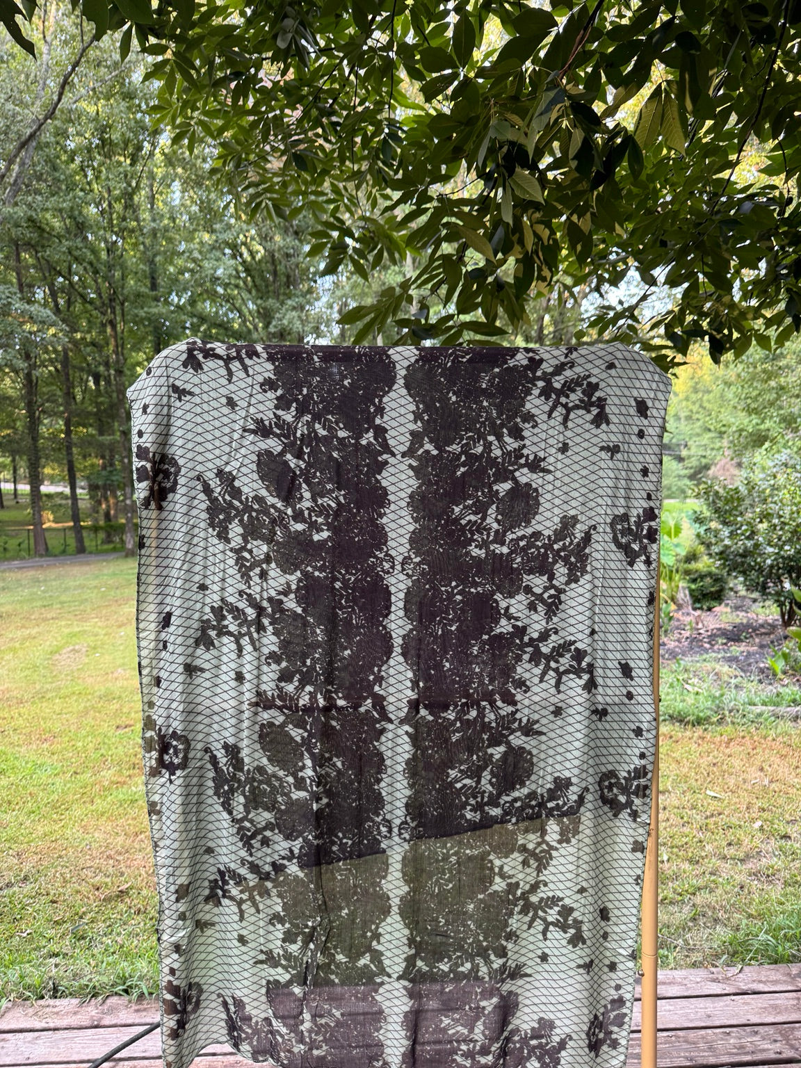 A lightweight patterned throw blanket with a black and off-white abstract design is draped over a wooden stand outdoors on a wooden deck, with trees and grass in the background.